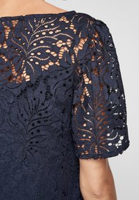 Top en dentelle bleu marine mettant en valeur des motifs de feuilles complexes, avec des manches courtes ballons et un tissu texturé avec des découpes sur les épaules.