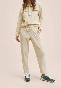 Beige Sweatshirt mit Kragen und Fronttasche, kombiniert mit passenden beige Jogginghosen mit seitlichen Paspeln, dazu grüne und weiße Sneakers.