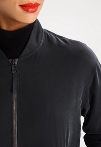 Chaqueta bomber negra con una textura suave, cuello alzado, cremallera frontal y un brillo sutil, combinada con un cuello de tortuga negro debajo.