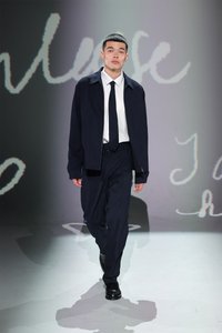 Marineblauwe jacket met rits aan de voorkant, witte shirt en donkere stropdas, gecombineerd met loszittende marineblauwe broek. Zwarte schoenen maken de outfit compleet. Eenvoudige achtergrond.