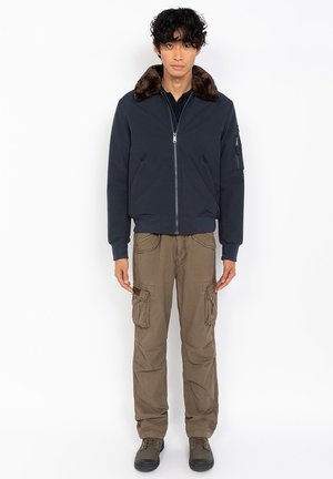 Giacca bomber blu con colletto in pelliccia marrone, chiusura con zip e tasche laterali; abbinata a pantaloni cargo beige e calzature scure.
