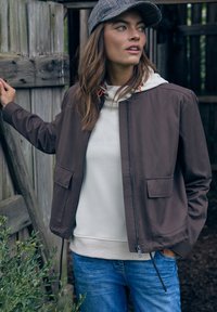 Femme portant une veste marron, un sweat à capuche beige, un jean bleu et une casquette à carreaux, debout en plein air près d'une clôture en bois, regardant sur le côté.