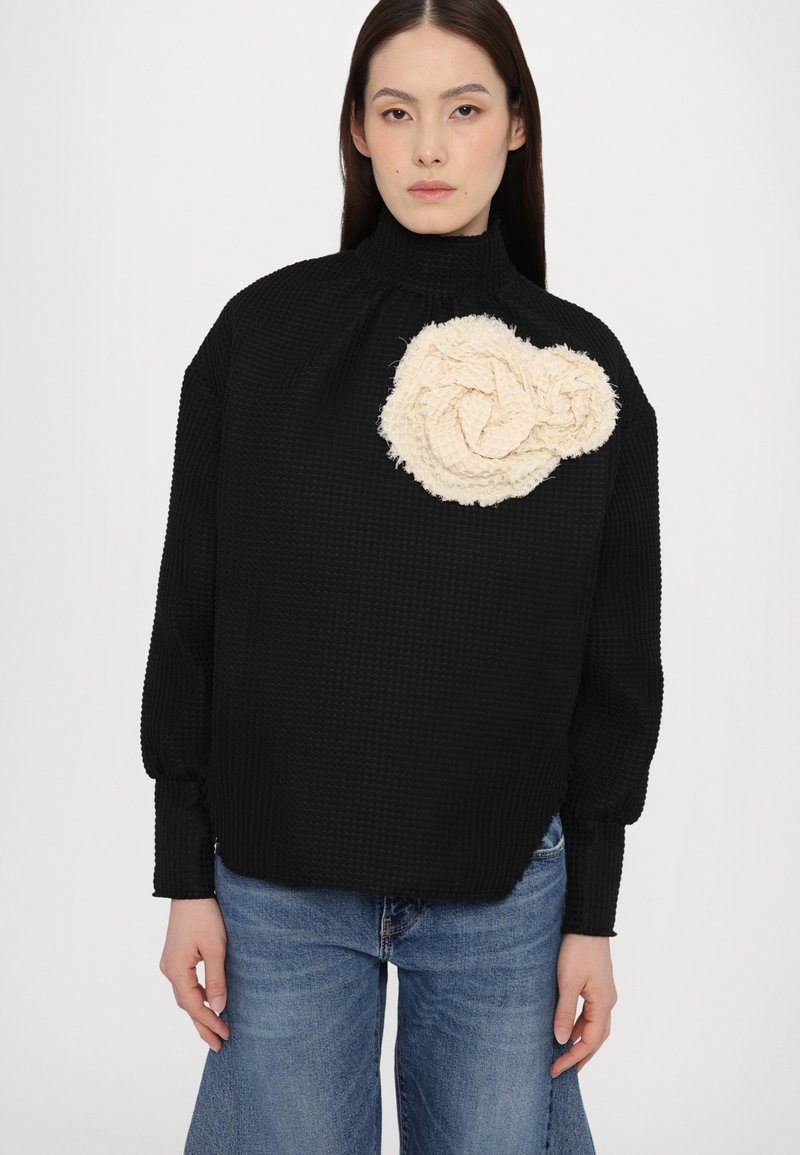 Sort tekstureret pullover med høj hals og løs pasform, der har en stor cremet knude detalje på venstre side. Blå denimbukser bæres.