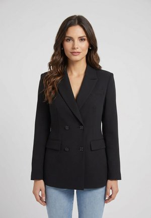 Mujer con cabello largo y ondulado de color marrón, vistiendo un blazer negro entallado de doble botonadura y vaqueros azul claro, de pie frente a un fondo liso.