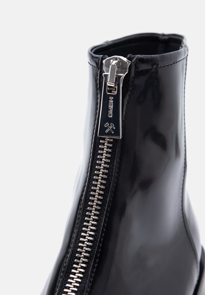 Μποτάκι αστραγάλου από μαύρο βερνικωμένο δέρμα με κεντρικό ασημένιο φερμουάρ που διαθέτει έλξη με χαραγμένο το "GMBH" και σύμβολα σφυριών.