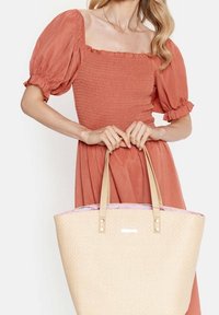 Borsa tote woven beige con interno rosa chiaro, due manici beige e accenti metallici. La borsa è tenuta da una persona che indossa un vestito color ruggine.