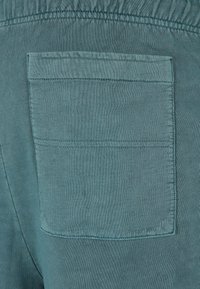 Teal stoff med en glatt tekstur, med en enkelt baklomme med en rektangulær klaff og horisontal søm. Elastisk midjebånddetalj.