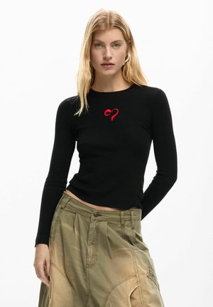 Czarna, prążkowana bluzka z długim rękawem z czerwonym haftem serca na piersi, zestawiona z luźnymi, khaki spodniami. Jasne, białe tło.