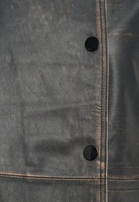 Primo piano di una giacca di pelle nera usurata con due bottoni a pressione neri lucidi e cuciture visibili lungo il bordo.