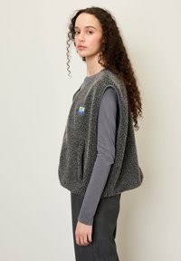 Mīksts pelēks villains veste ar lielām roku atverēm, sānu kabatām un mazu izšūtu plāksteri; valkāta virs pieguļošas pelēkas kreklu ar garām piedurknēm.