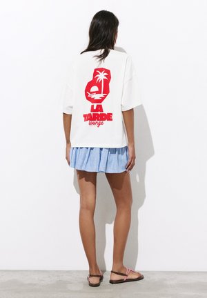 Femme debout face à un mur blanc portant un t-shirt blanc oversize avec un motif de palmier rouge, une jupe rayée bleue et des sandales.