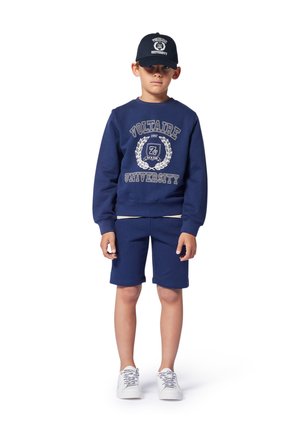 Sweat-shirt bleu marine avec le logo "UNIVERSITÉ VOLTAIRE", shorts assortis et baskets blanches ; une casquette bleue marine vient compléter la tenue.