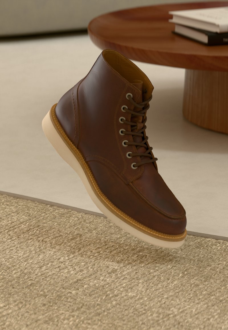 Bottine en cuir marron avec semelle beige sur un tapis beige texturé à côté d'une table ronde en bois avec des livres empilés.