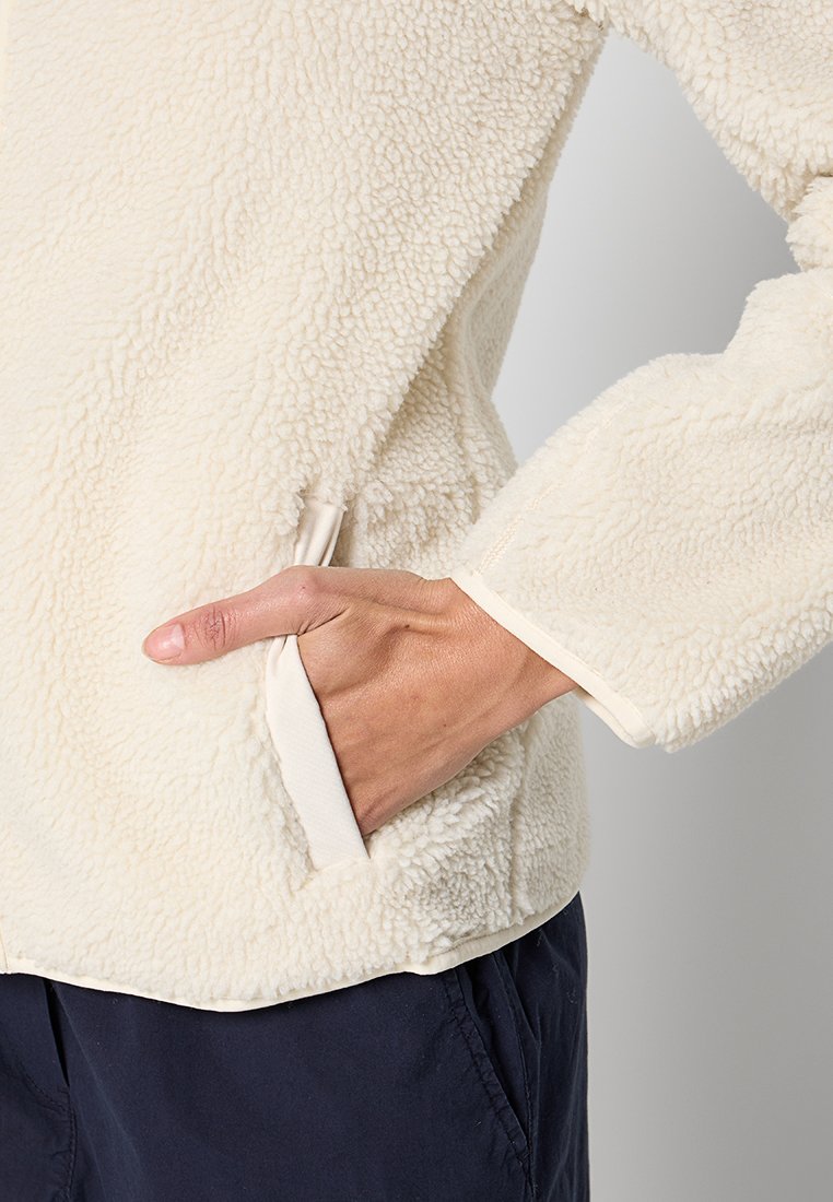 Main dans une poche de veste en polaire beige, montrant une texture douce et un ajustement décontracté sur un pantalon sombre, sur un fond neutre.