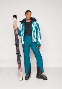 Hiiemeskidress, millel on sinine ja valge mustriga jope, must pehme pluus, teal värvi püksid ja mustad suusasaapad, hoides ühes käes suusakiudu koos sidemega.