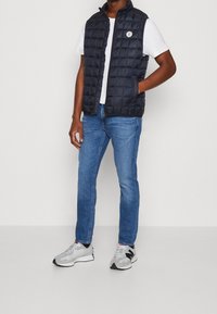 Marinblå puffväst med dragkedja fram, ståkrage och sidofickor, parat med en vit t-shirt, blå jeans och grå sneakers.