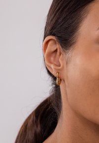 Goldene Creolen mit einem wellenförmigen Design, verziert mit klaren Strasssteinen. Am Ohr getragen, betonen sie das Ohrläppchen und die nach hinten gebundenen Haare.