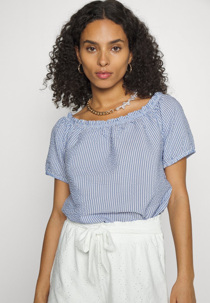 Blusa às riscas azuis e brancas com mangas curtas, um decote franzido e um corte relaxado, combinada com uma saia branca texturizada.