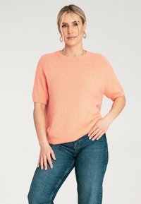 Krótki rękaw, jasnoróżowy sweter wykonany z miękkiego, puszystego materiału, z okrągłym dekoltem i luźnym krojem, w połączeniu z niebieskimi dżinsami.