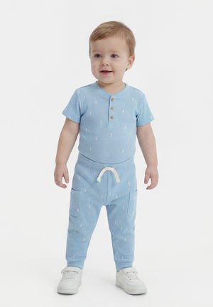 Kisgyermek áll és mosolyog, világoskék rövid ujjú felsőt és hozzáillő nadrágot visel fehér sportcipőkkel, fehér háttér előtt.