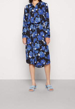 Femme portant une robe midi noire à imprimé floral bleu, manches longues et ceinture nouée à la taille, associée à des sandales à talons bleu clair.