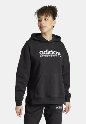 Giovane con capelli ricci indossa felpa nera con cappuccio Adidas e pantaloni abbinati con loghi bianchi Adidas Sportswear sulla parte frontale.