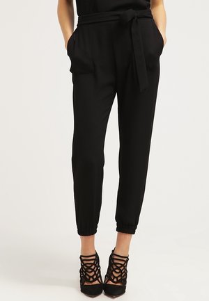 Femme portant un pantalon noir taille haute à coupe fuselée avec ceinture nouée à la taille et chaussures à talons hauts noires à brides, les mains dans les poches.