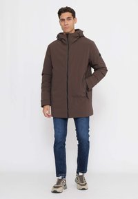 Braune Jacke mit Reißverschluss und Kapuze aus glattem Stoff, mit zwei Seitentaschen und schwarzen Akzenten. Kombiniert mit blauen Jeans und Sneakern.
