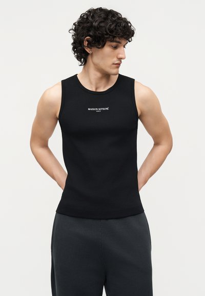 Jeune homme aux cheveux bouclés portant un débardeur noir sans manches avec le logo "Maison Kitsuné Paris", mains derrière le dos, regardant vers sa gauche.