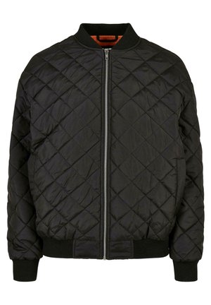Femme portant une veste bomber matelassée noire, un haut noir, un pantalon large blanc taille haute et des baskets noires, debout devant un fond blanc.