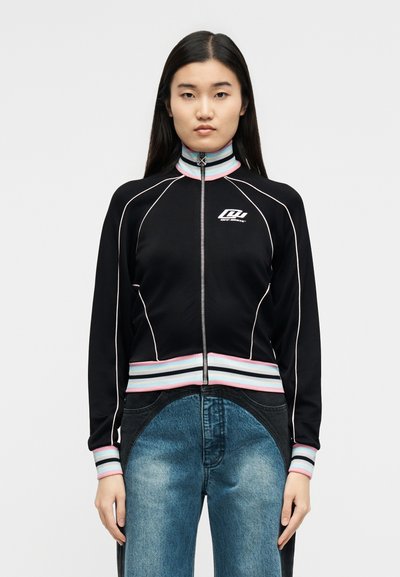 Νεαρή γυναίκα με μακριά σκούρα μαλλιά, ντυμένη με μαύρο μπουφάν με φερμουάρ, λευκές και παστέλ λεπτομέρειες, και μπλε τζιν με δύο αποχρώσεις, στέκεται μπροστά σε απλό φόντο.