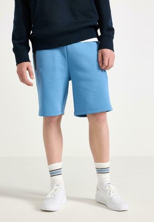 Person iført lyseblå knælange shorts, hvide atletiske sokker med blå striber, hvide sneakers og en sort sweatshirt.