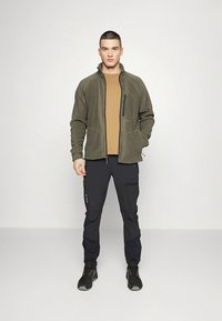 Olíva zöld polárdzseki cipzáras zsebbel, barna ing alatt, fekete nadrág, fekete túracipő. Egyszínű háttér, elölnézet.