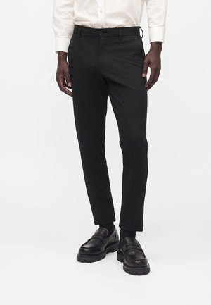 Muž v čiernych slim fit nohaviciach, čiernych mokasínach a bielej košeli na gombíky, stojaci pred jednoduchým bielym pozadím.