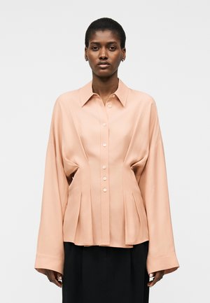 Γυναίκα που φοράει φαρδιά, ανοιχτόχρωμη ροδακινί μπλούζα με κουμπιά και φαρδιά μακριά μανίκια, καθώς και μαύρο παντελόνι, στέκεται μπροστά σε απλό φόντο.