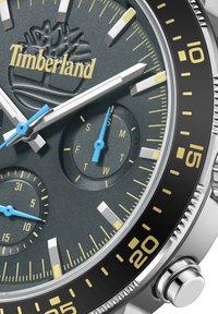Hodinky Timberland s textúrovaným šedým ciferníkom, striebornými markerami, modrými ručičkami a čiernym rámom s časovými indikátormi a subciferníkom na deň/dátum.