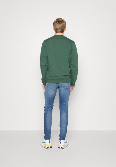 Grüne Sweatshirt mit rundem Halsausschnitt und gerippten Bündchen, kombiniert mit schmalen blauen Jeans und weißen Sneakers mit gelben und blauen Akzenten.