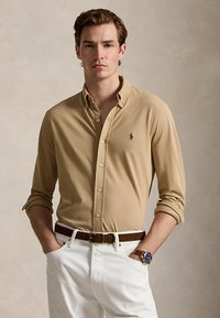 Jeune homme portant une chemise beige boutonnée avec un petit logo, un pantalon blanc, une ceinture en cuir marron et une montre-bracelet au cadran bleu, debout les mains dans les poches.