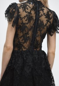 Γυναίκα που φοράει μαύρο φόρεμα από δαντέλα με λουλουδάτα σχέδια και διαφανή πλάτη, με ζώνη στο κέντρο και κοντά μπαλονέ μανίκια.