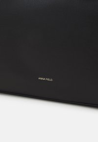 Surface en cuir noir texturé avec un petit logo de marque doré "ANNA FIELD" centré près du bord inférieur.