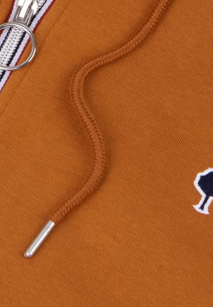 Particolare di un tessuto arancione con un cordino intrecciato, agglomerato metallico, zip argentata e un piccolo logo di un albero ricamato in bianco e nero.