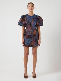 Donna in piedi che indossa un vestito corto multicolore con maniche a palloncino e vita aderente, con tacchi a punta marroni su uno sfondo semplice.