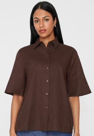 Frau mit langen schwarzen Haaren trägt ein braunes Hemd mit kurzen Ärmeln und Kragen, steht vor einem einfarbigen hellen Hintergrund.