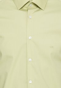 Camicia verde chiaro con bottoni, realizzata in tessuto liscio, con colletto appuntito e bottoni bianchi, con un sottile logo del marchio ricamato sul petto.