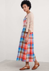 Maxikleid mit mehrfarbigem Karomuster in Rot, Blau und Beige. Kombiniert mit einer hellbeigen Strickjacke und braunen gewebten Slippern.