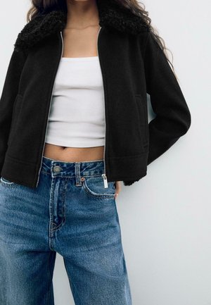 Vrouw draagt een zwarte jas met rits en een gestructureerde kraag over een wit cropped topje en hoge taille blauwe spijkerbroek.