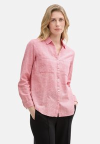 Camicia a maniche lunghe rosa chiaro in tessuto testurizzato con bottoni anteriori, colletto e due tasche sul petto; abbinata a pantaloni neri.