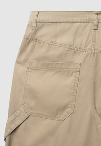 Nahaufnahme von beigen Baumwollhosen, die die Gesäßtasche, Gürtelschlaufen und seitliche Riemen_details mit klaren Nähten auf einem einfarbigen Hintergrund zeigen.