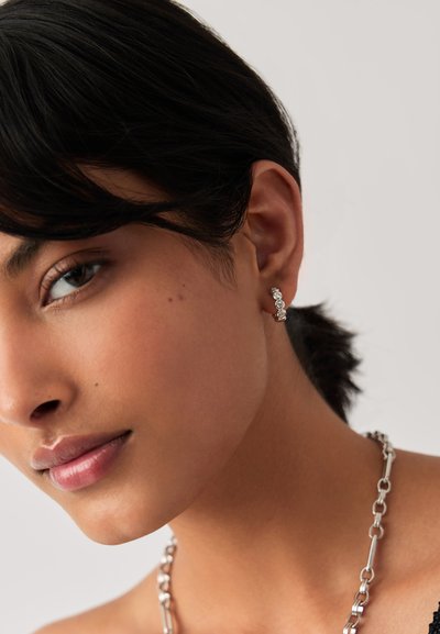 Κοντινό πλάνο ενός ατόμου με κοντά σκούρα μαλλιά, που φοράει ασημένιο κολιέ με αλυσίδα και ασημένιο σκουλαρίκι με τρεις μικρές πέτρες.