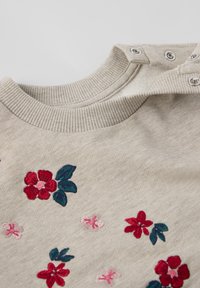 Lichtbeige sweatshirt met geborduurde bloemen in rood en roze, voorzien van een ribgebreide kraag en drukknopen op de schouder. Zachte textuur.
